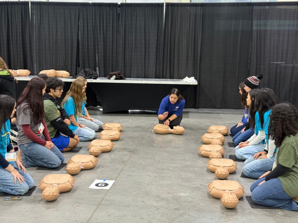 A group of students sitting on a gray floor learning about Hands-Only CPR from an instructor