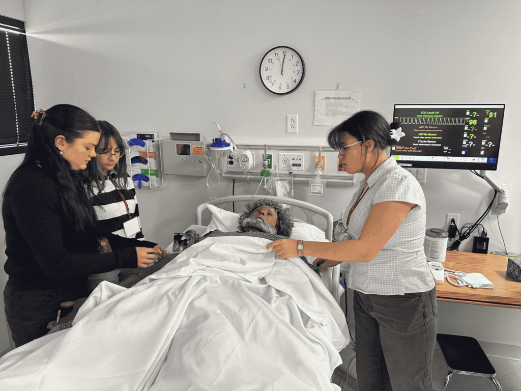 Three female students stand around a hospital bed in a simulated hospital room 