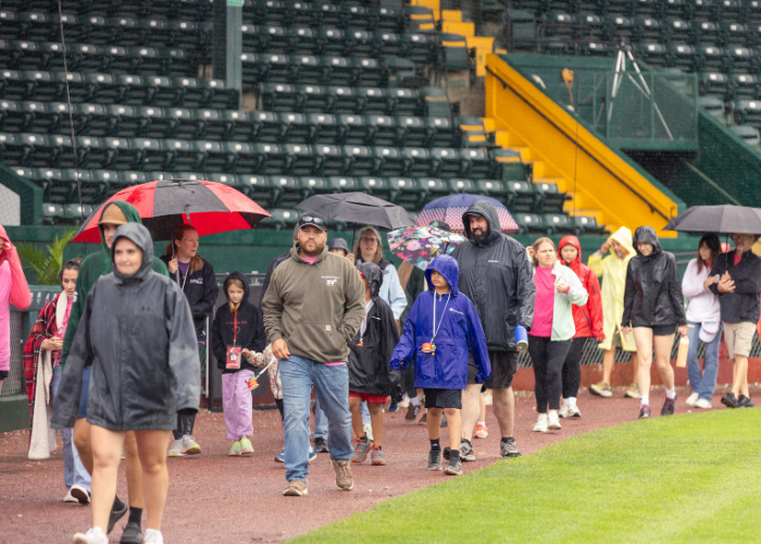 Crowd of people walking at baseball field with umbrellas.