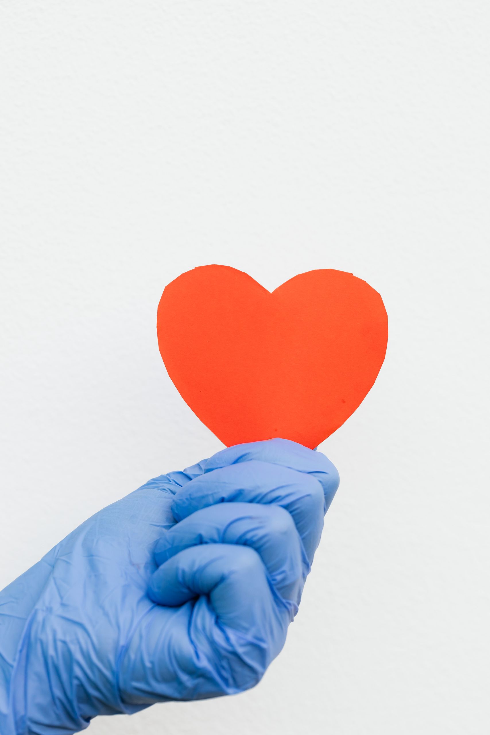 A blue gloved hand holding a red paper heart against a white background, symbolizing care and compassion.