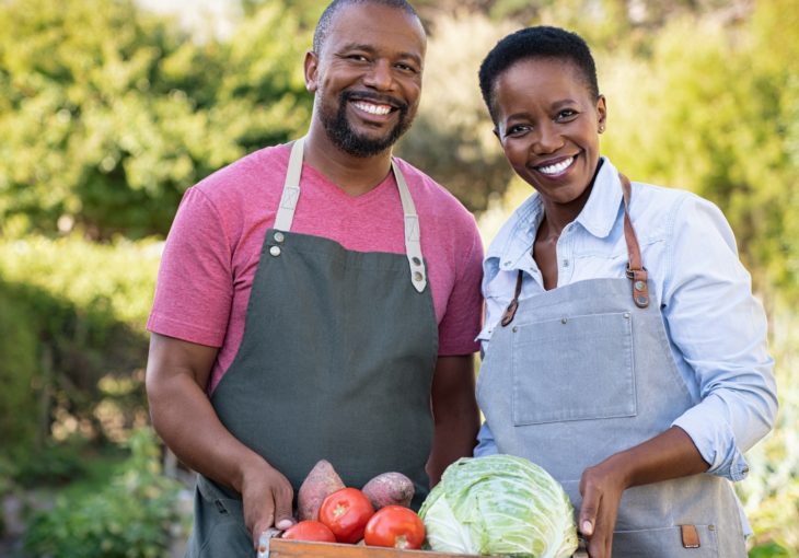 African-American man and woman standing in a field holding box of vegetables.
