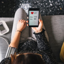 Young woman measures blood pressure sitting on sofa at home with smartphone connected to device – Concept of health, well-being and love for oneself – Millennial in a moment of private life
